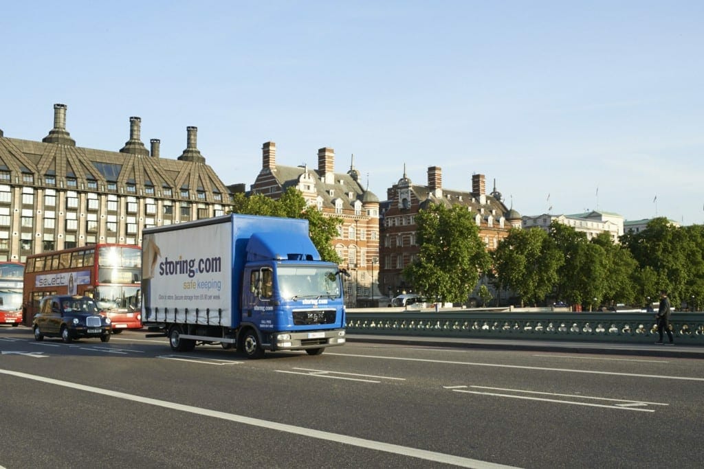 A blue self storage company truck driving over a bridge with city buildings in the background under a clear sky.