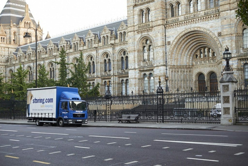 A blue delivery truck labeled "strong.com" drives past the ornate facade of the natural history museum in London, advertising self storage solutions.