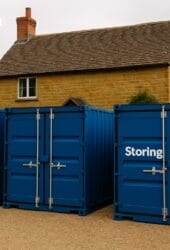 Three blue storage containers labeled "Storing.com" are placed in front of a house with stone walls and a tiled roof. The word "Biddenham" appears in the top left corner.